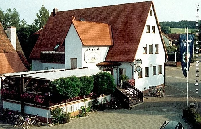 Ein großes vierstöckiges Gebäude mit weißer Fassade und rotem Dach. Links an das Gebäude ist eine lange überdachte Terasse aus Holz angebaut.