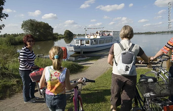Eine Familie läuft mit ihren Fahrrädern auf eine Anlegestelle zu. Dort wartet ein Schiff.