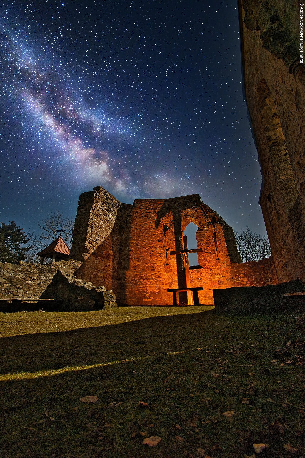 Die Katharinenkapelle bei Nacht. Indirket beleuchtet durch orange gelbe Lichter. Der Sternenhimmel blau lila und funkelt.