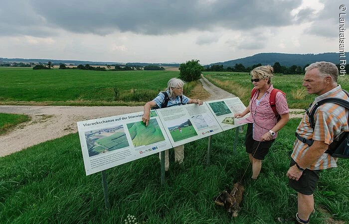 Drei Wanderer stehen um eine Infotafeln in der grünen Landschaft. Rund herum grüne Wiesen.