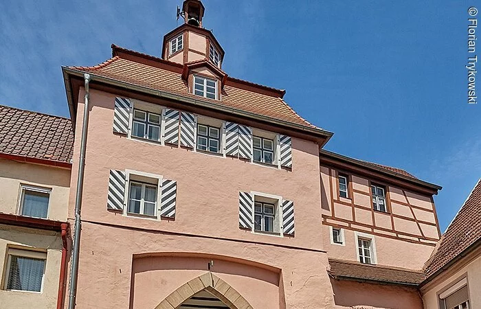 Vier Radfahrer radeln über eine geplasterte Straße durch ein Stadttor. Das Stadttor hat eine rosane Fassade und blau-weiß gestreifte Fensterläden.
