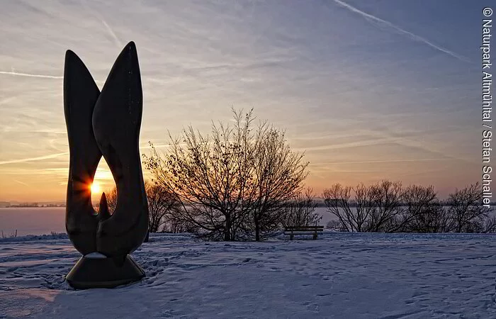 Kunstobjekt bei Sonnenaufgang am schneebedecktem Altmühlsee. Einige Meter dahinter stehen einige Sträucher.