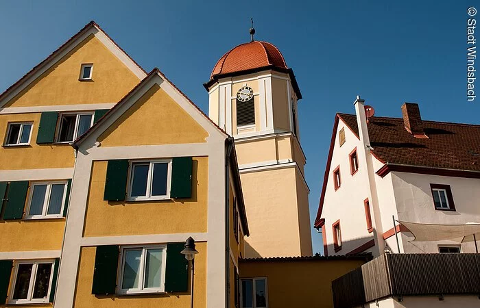 Blick von unten auf eine Kirchturmspitze. Ein Turm mit weiß-gelber Fassade und rotem Haubendach. Davor stehen Häuser im fränkischen Baustil. Links ein gelbes Haus mit grünen Fensterläden. Rechts ein Haus mit weißer Fassade.