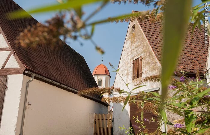 Ausschnitt von zwei älteren Häusern. Zwischen den Häusern ragt ein Kirchturm hervor. Der Turm hat eine weiß-gelbe Fassade mit rotem Haubendach.