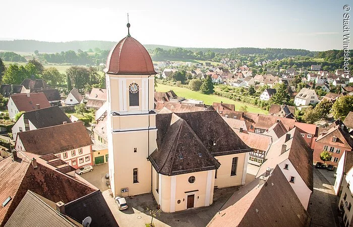 Luftbild einer Kirche umgeben von Häusern. Ein Kirchenschiff und Kirchturm mit heller oragener Fassade. Der Kirchturm hat ein Haubendach.