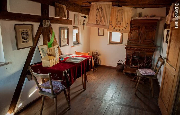 Blick in eine alte bürgerliche Stube. Fachwerkwand mit weißen Wänden. Holzboden und Holzmöbel. In der rechten Ecke ein alter Holzschrank. Links ein Tisch mit zwei Stühlen.