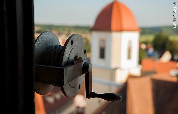Im Fokus eine Spule mit Seil und Kurbel. Im Hintergund ist verschwommen ein Turm zu erkennen. Der Turm hat eine helle gelbe Fassade und ein rotes Haubendach.