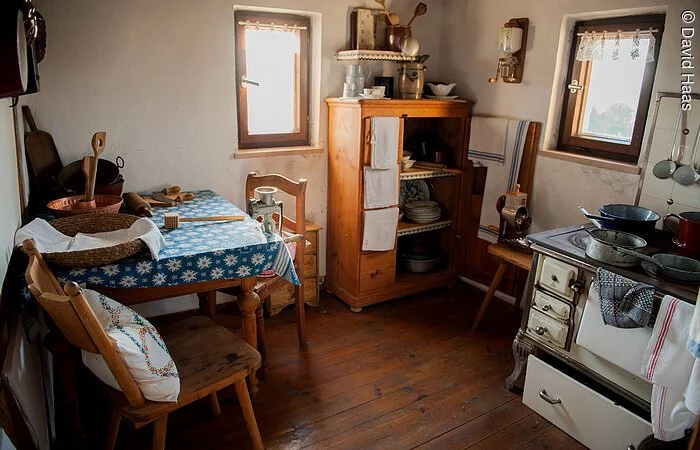 Eine kleine altertümliche Küche. Sitzecke mit zwei Stühlen in der rechten Ecke. in der hinteren Ecke ein Holzschrank. Rechts davon ein alter Holzofen zum Kochen.