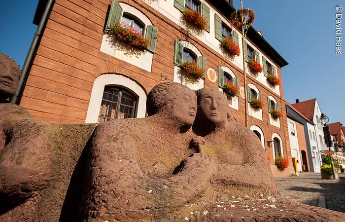 Blick und untern auf zwei Figuren eines Steinbrunnen. Die zwei Figuren halten Hände. Im Hintergund ein großes Haus mit Steinfassade. An den Fenstern grüne Fensterläden und Blumenkästen mit roten Blumen.