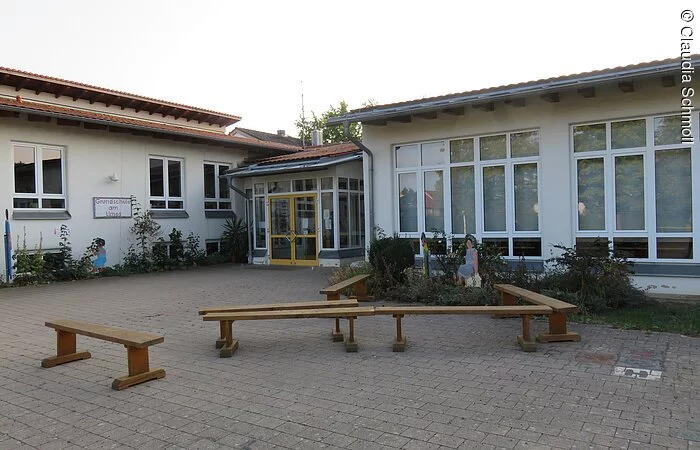 Außenansicht eines ebenen Hauses mit hohen Fnstern. Weiße Fassade und rotes Ziegeldach. Davor ein gepflasteter Hof mit fünf Sitzbänken aus Holz.