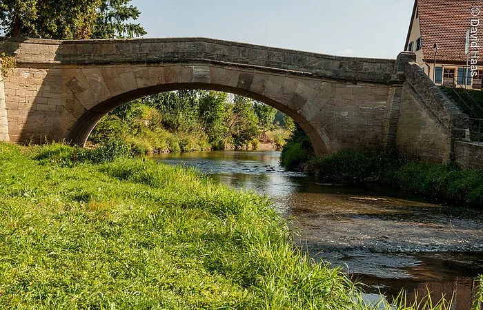 Eine Steinbrücke führt über einen Fluss. HInter der Brücke wachsen am Ufer Bäume. Am Ufer vor der Brücke eine grüne Wiese.