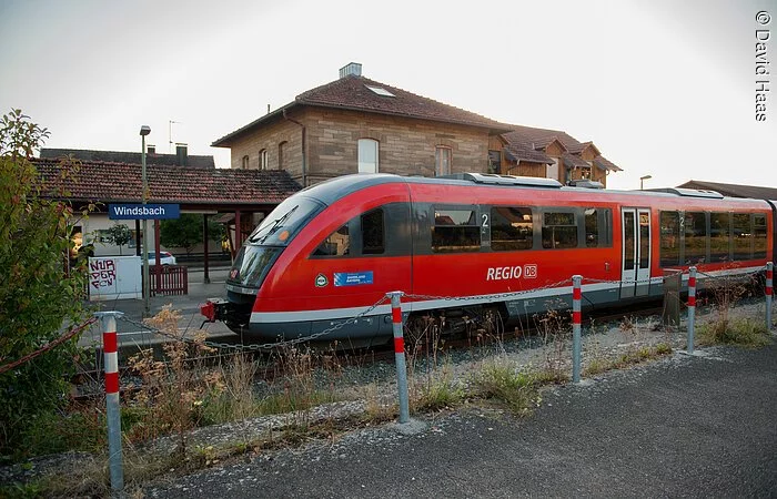 Ein kleiner Bahnhof. Auf dem einzigen Gleis steht ein roter Zug der deutschen Bahn. Dahinter ein Haus mit Steinfassade.