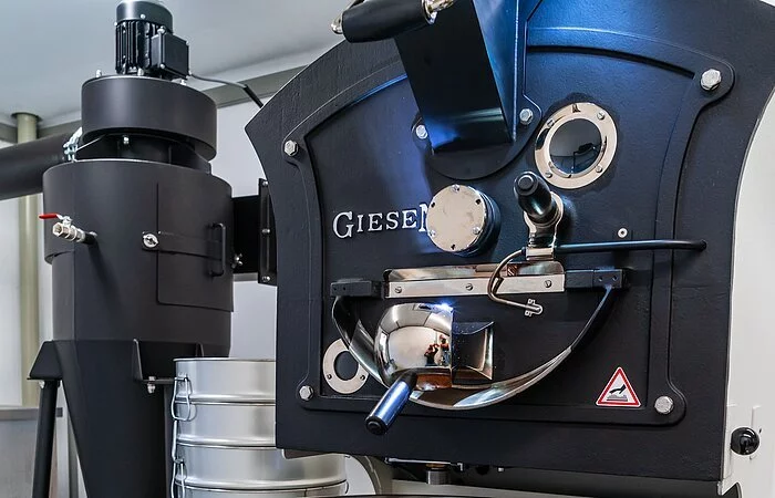 Eine Kafferöstmaschine. Eine große weiße Blechtrommel mit einem Rüherer, der die Kaffeebohnen in der Trommel rührt.