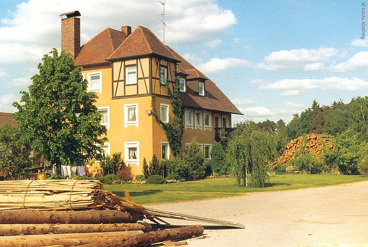 Foto eines Wohnhauses mit drei Ebenen. Links ein Baum und Davor liegen Holzstämme auf einem Haufen.