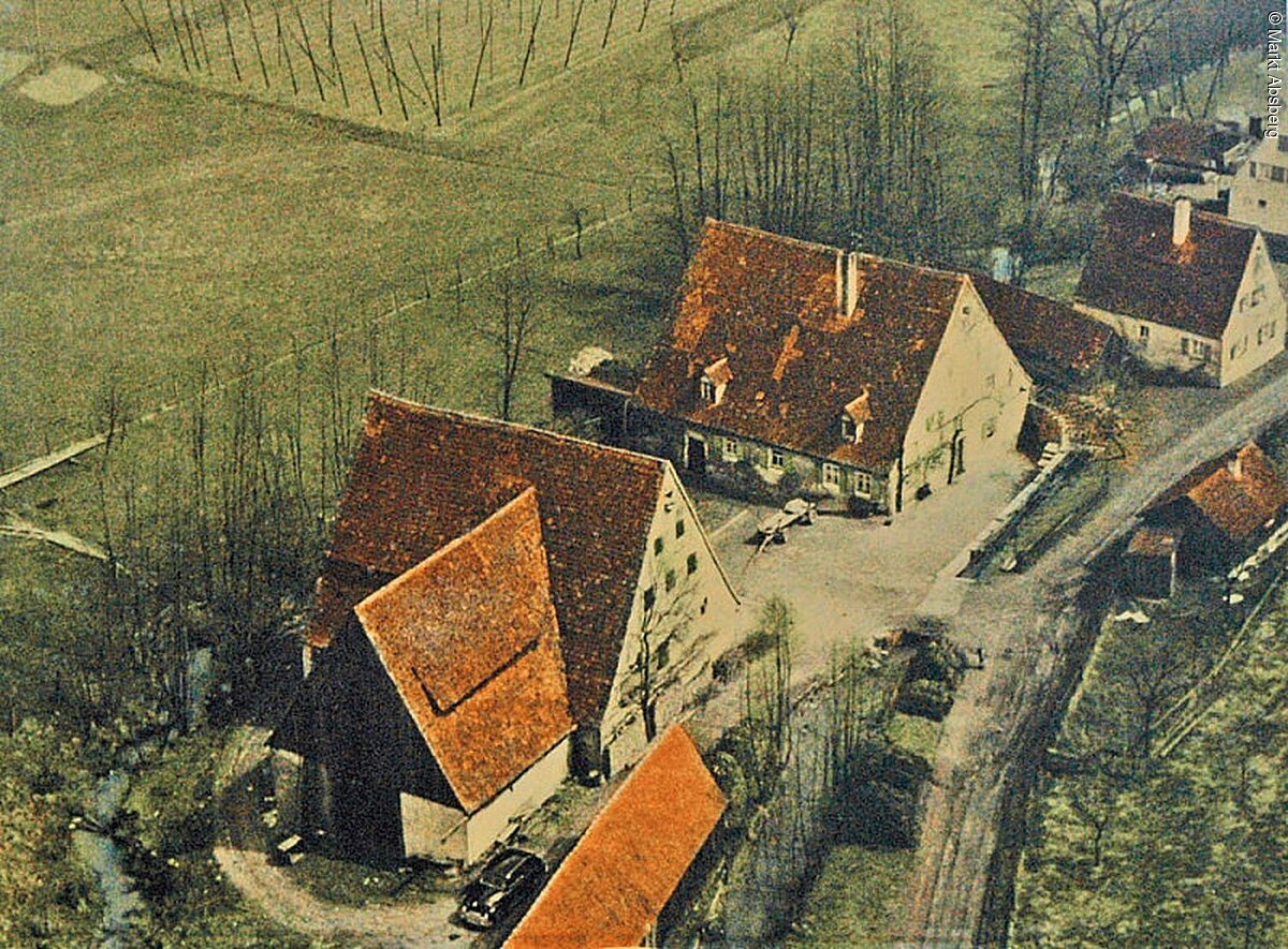 Ein altes Foto einer Mühle. Drei Häuser nebeneinander. Vorne vorbei führt ein Weg. Dahinter sind Felder.