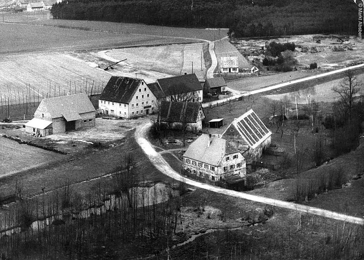 Ein schwarz-weiß Foto eines Mühlenkomplexes. Vier alte Häuser auf einem Grundstück. Durch das grundstück führt rin Weg. Drum herum Wiesen und Wälder.