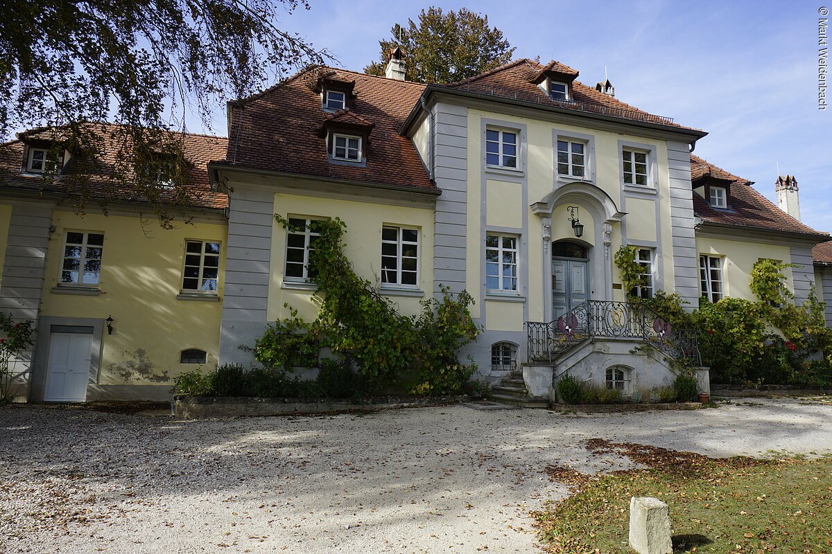 Ein barockes Gebäude mit gelber Fassade und rotem Ziegeldach. Das Haus hat mehere Unterteilungen. Am Eingang eine große Treppe die rechts und links von der Fassade hinauf zum Eingang führt.