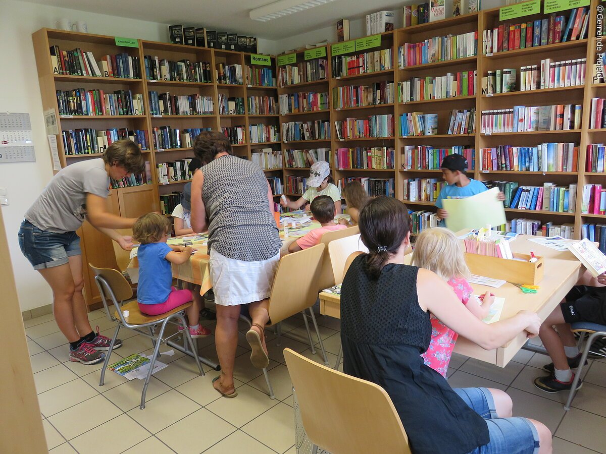 Eine Bücherei mit gefüllten Bücherregalen. Kinder und Erwachsene sitzen an Tischen.