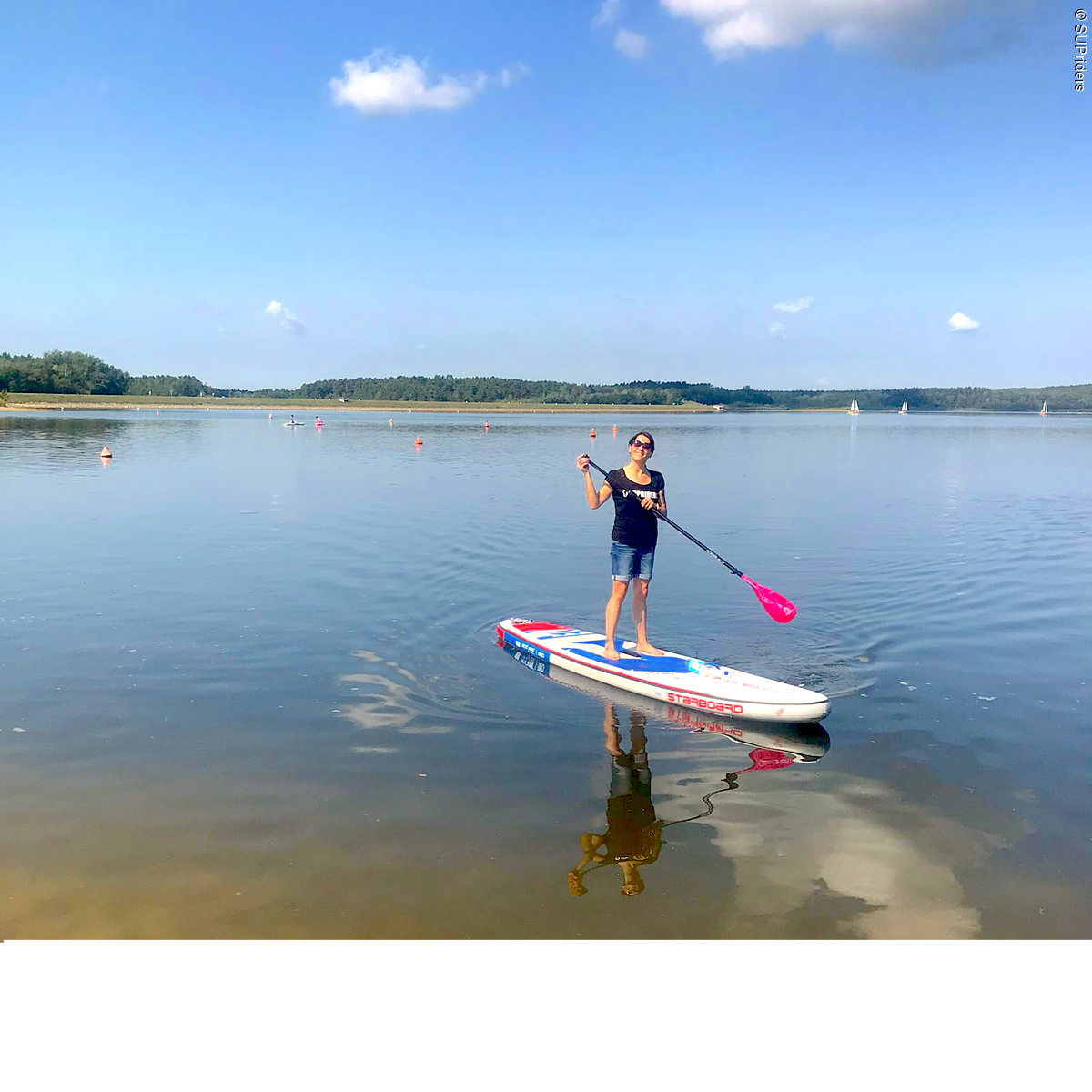 Eine junge Frau auf einem Stand Up Paddle mitten auf dem See. Der blaue Himmel spiegelt sich im Wasser.