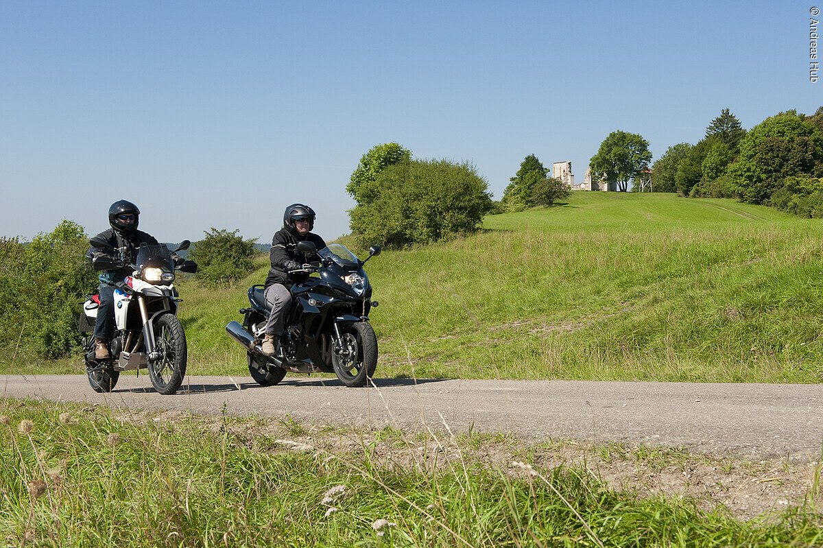 Zwei Motorradfahrer fahren auf einer geteereten Straße durch die grüne Landschaft des Fränkischen Seenlands. Im Hintergrund ist eine grüne Erhebung sowie eine Baumreihe zu erkennen.