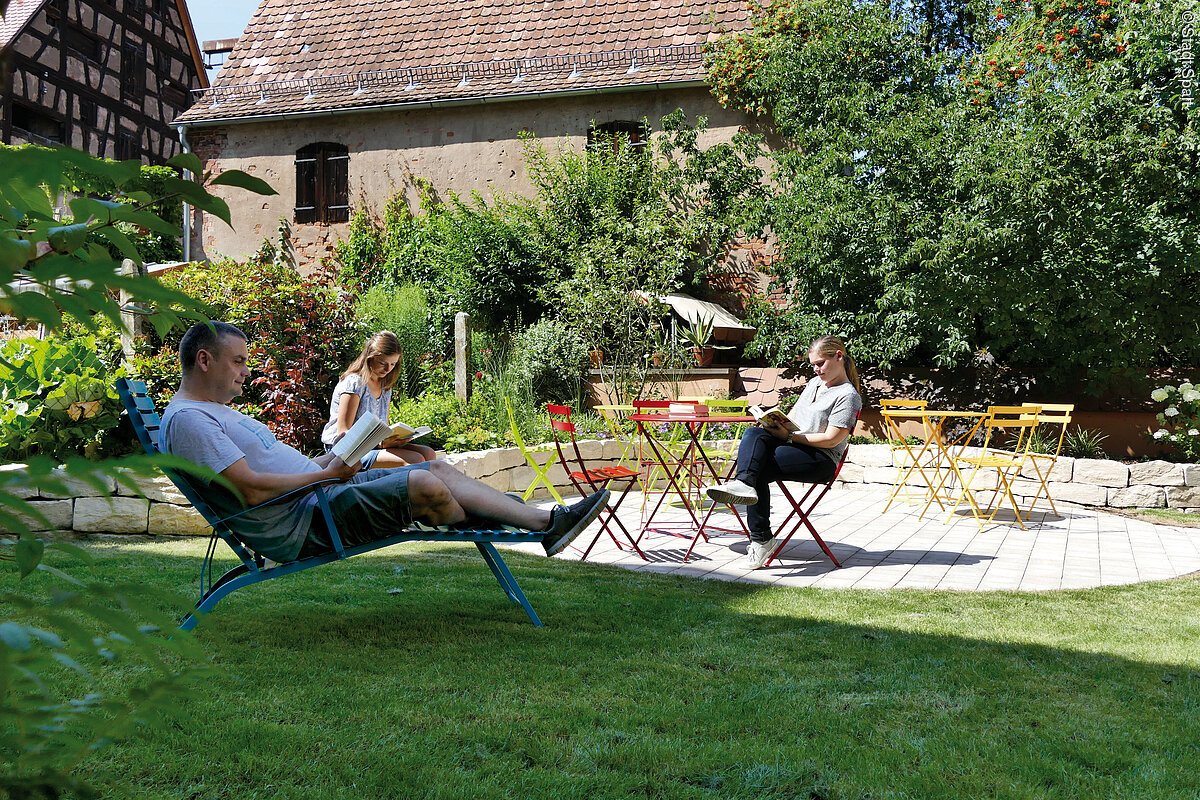 Ein grüner Garten mit Wiese und Sträuchern. Ein Mann und zwei junge Frauen sitzen im Garten und lesen Bücher.