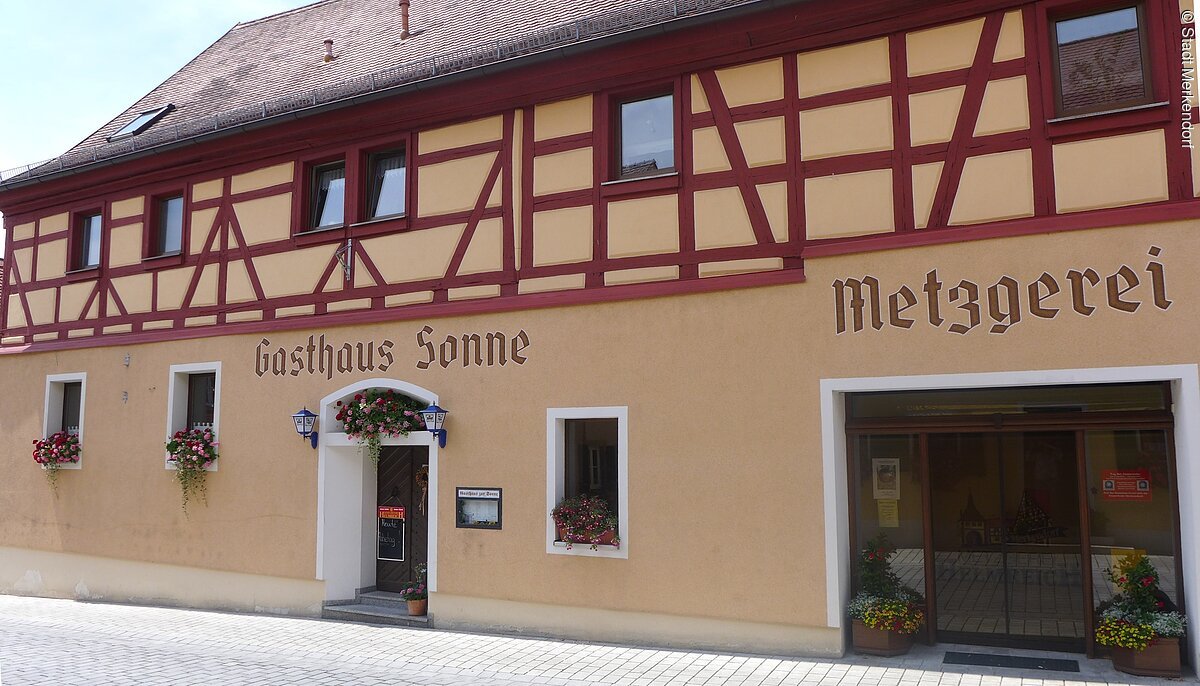 Frontansicht des Gasthaus Sonne mit angrenzender Metzgerei auf der rechten Seite. Ein großes hell-orangenes Fachwerkhaus mit roten Balken.