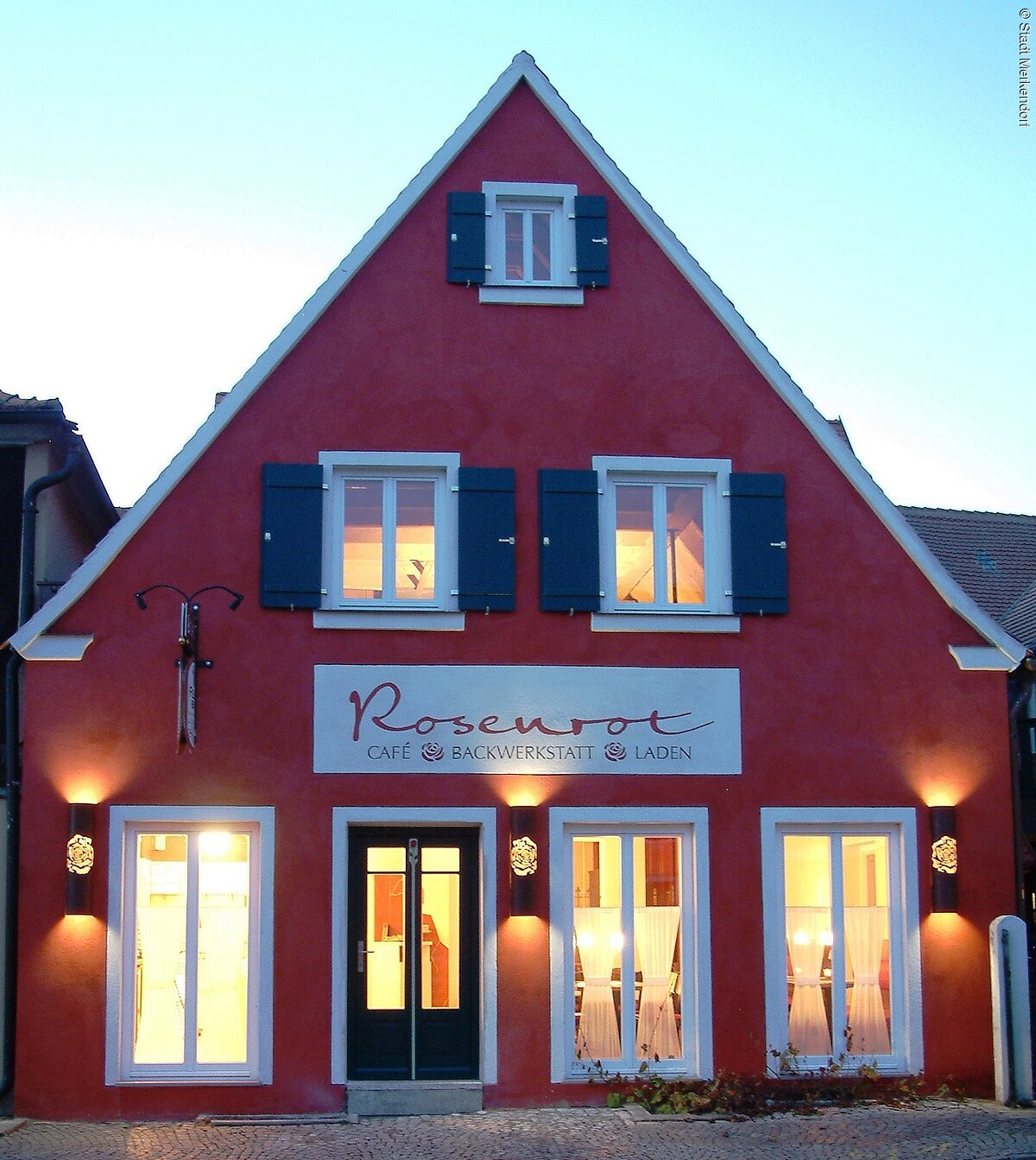 Die rote Außenfassade des Café Rosenrot mit blauen Fensterläden und  hellerleuchteten Fenstern in der Abenddämmerung.