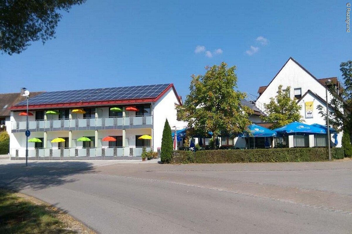 Ein langes zweistöckiges Gebäude mit weißer Fassade und Balkonen. Auf dem Dach eine  Solaranlage. Rechts daneben ein kleiner Biergarten mit Hecke und blauen Sonnenschrimen. Dahinter spitzelt ein weiteres Haus mit weißer Fassade hervor.