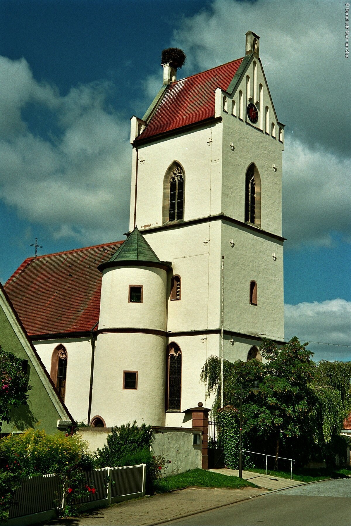 Ein Kirche mit weißer Fassade. Neben dem Kirchenschiff ein großer Kirchturm mit rotem Dach und Storchennest auf dem Schlot.