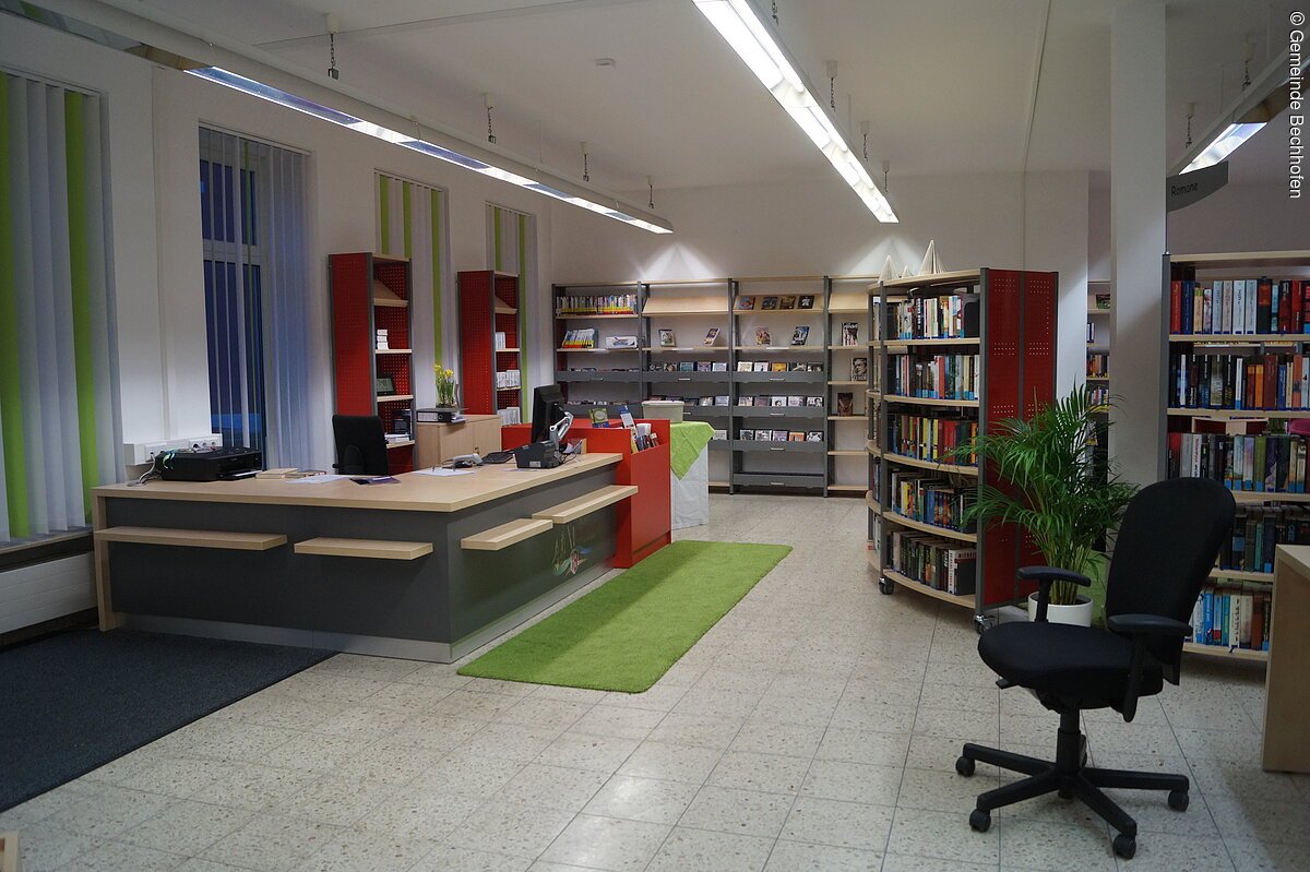 Eine Bücherei mit Empfangstresen auf der linken Seite. Dahinter rote Bücherregale. Links davon weitere Bücherregale.
