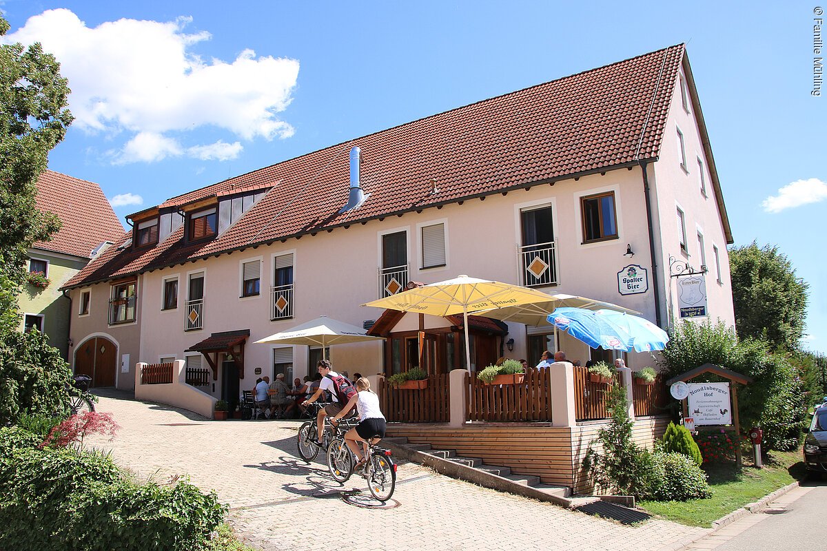Seitansicht der Gastwirtschaft Noudlsberger Hof. Ein rosa farbenes Haus mit einer Terasse mit Sitzgelegenheiten und Sonnenschirmen direkt davor. Zwei Radler radeln die Einfahrt des Hofes hoch.