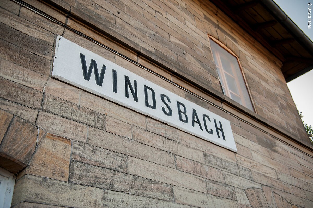 Nahaufnahme einer Hausfassade. An der Steinfassade hängt ein weißes Schild. Darauf in schwarzen Blockbuchstaben das Wort Windsbach.