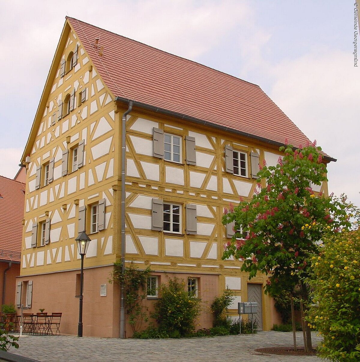 Ein Fachwerkhaus mit weißer Fassade und gelben Balken. Die Fensterläden sind grau. Da Dach aus roten Dachziegeln. Das Haus hat fünf Geschosse.