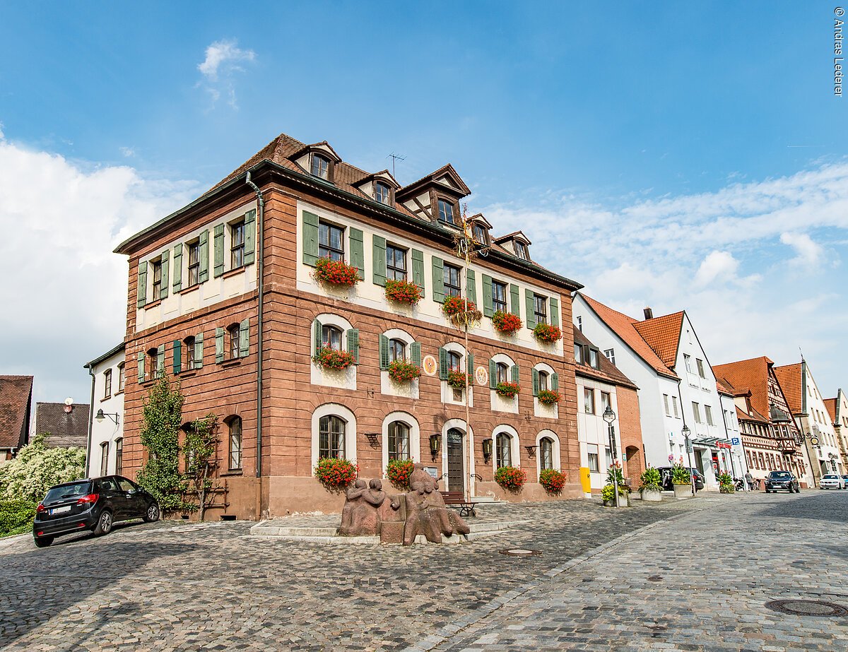 Ein großes Haus mit vier Ebenen. Ein Haus mit Steinfassade. An den Fenstern grüne Fensterläden. Daran Blumenkästen mit roten Blumen. Das Haus hat ein Walmdach. Das Haus liegt an einer gepflasterten Straße.