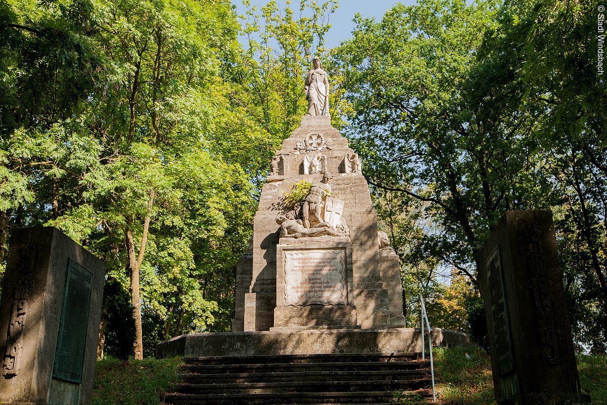 Ein Kriegerdenkmal umegben von Bäumen. Eine Treppe führt zu einem Steinsockel auf welchem ein Figurenensemble steht. Ein Mann liegt am Boden, ein weitere steht drüber und hält ein Schild. Einen Sockel weiter oben eine Frau mit langem Gewand.