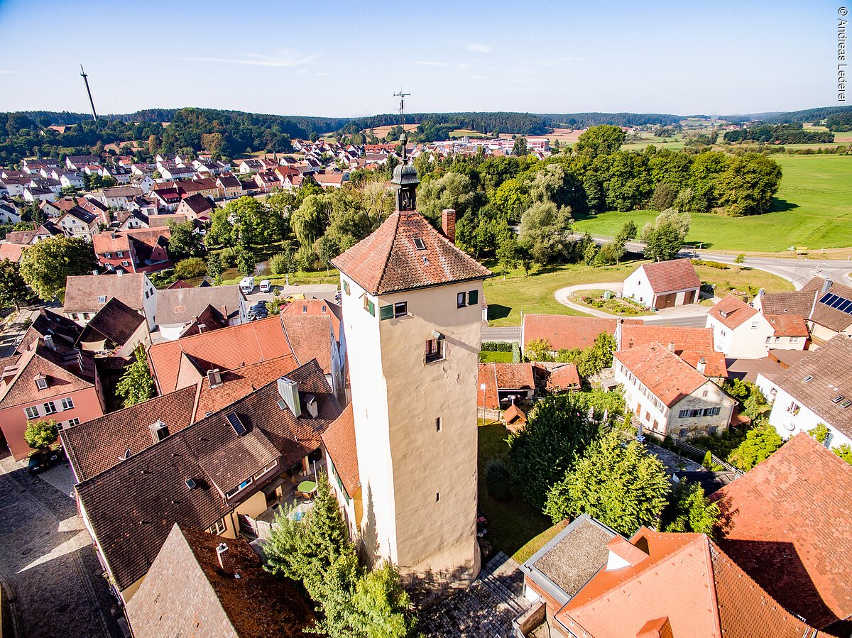Luftbild  eines Stadtturms. Ein großer Turm mit beiger Fassade und rotem Ziegeldach. Um den Turm Häuserdächer.