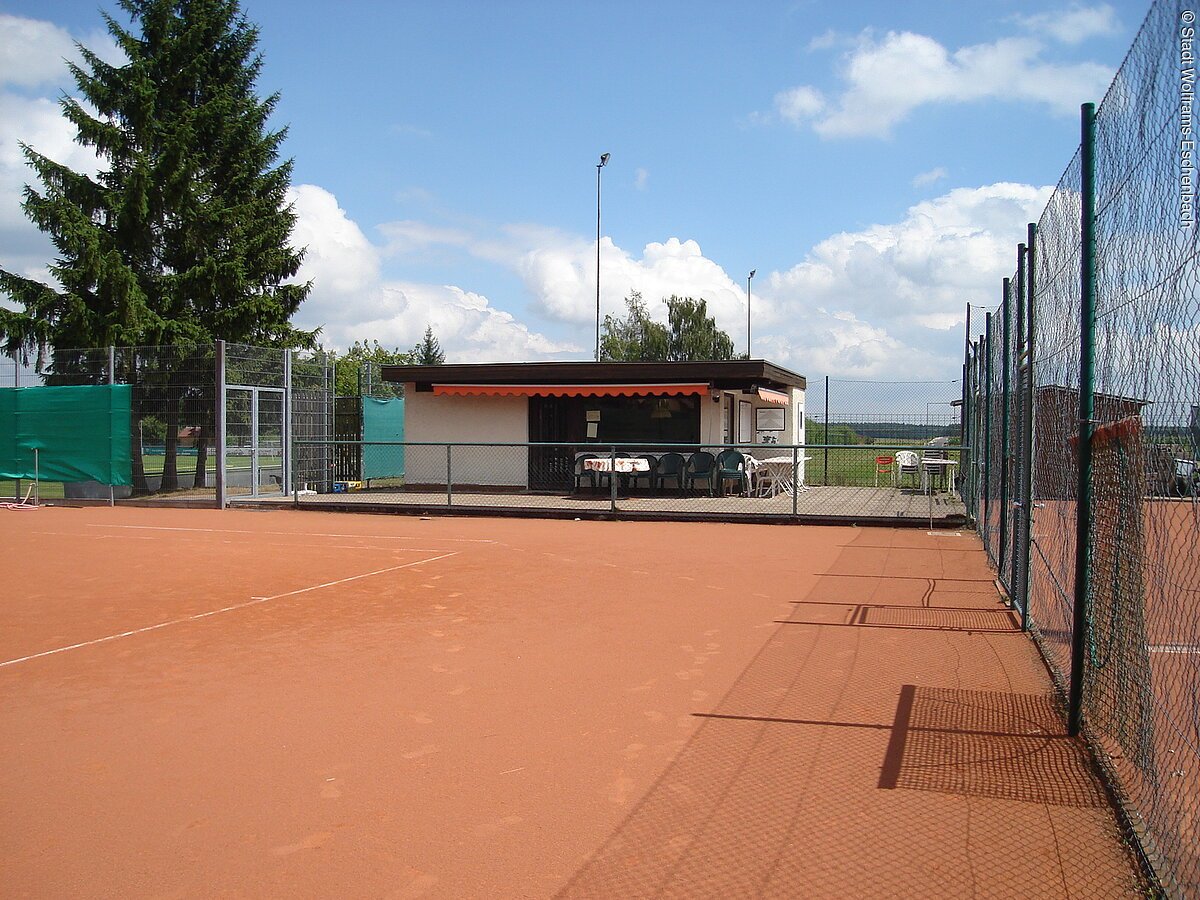 Ein roter Tennisplatz umrahmt von grünen Bäumen unter blauem Himmel.