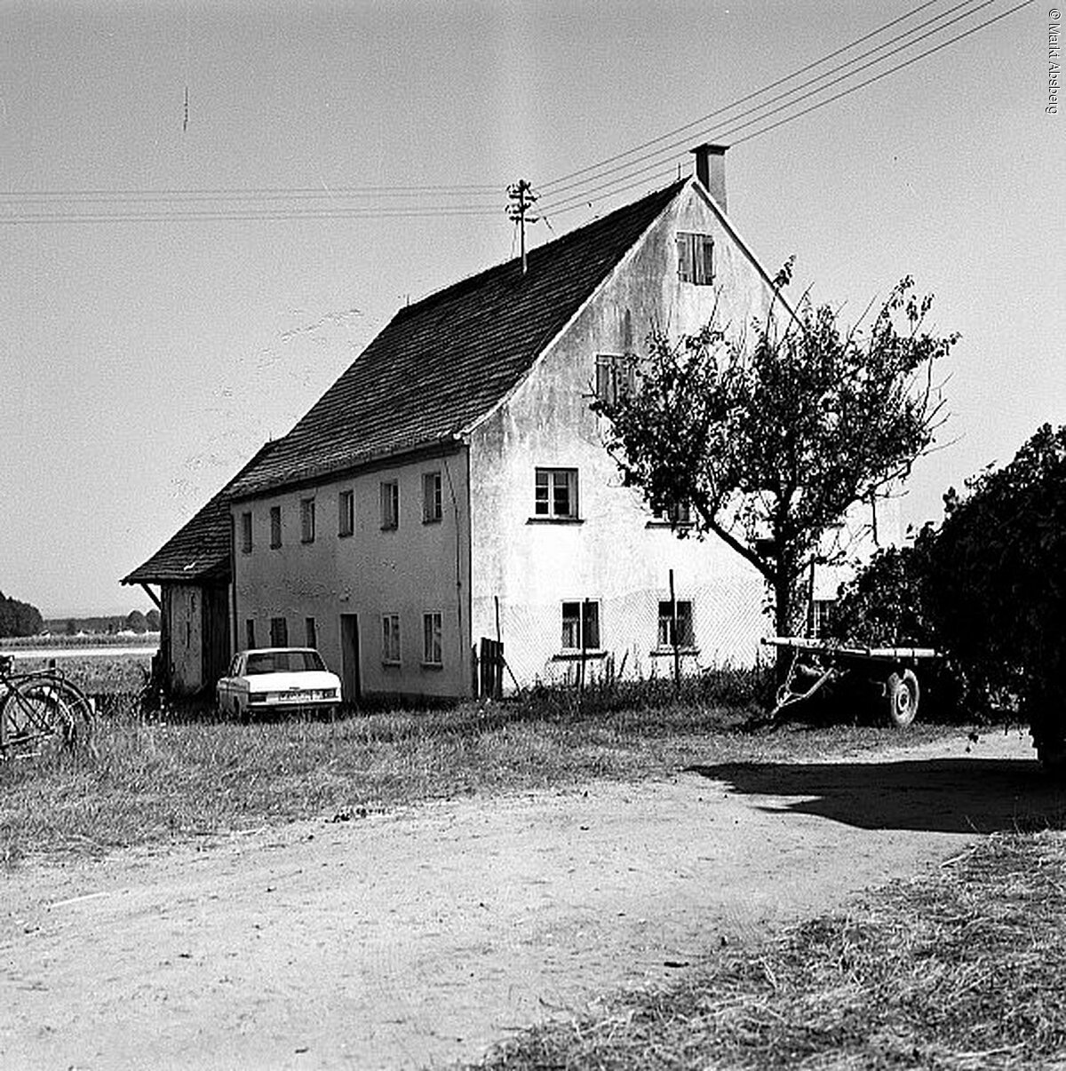 Schwarz-weiß Bild eines alten Hauses mit vier Ebenen. Vor dem Haus steht ein Baum. Links davon ein Auto.