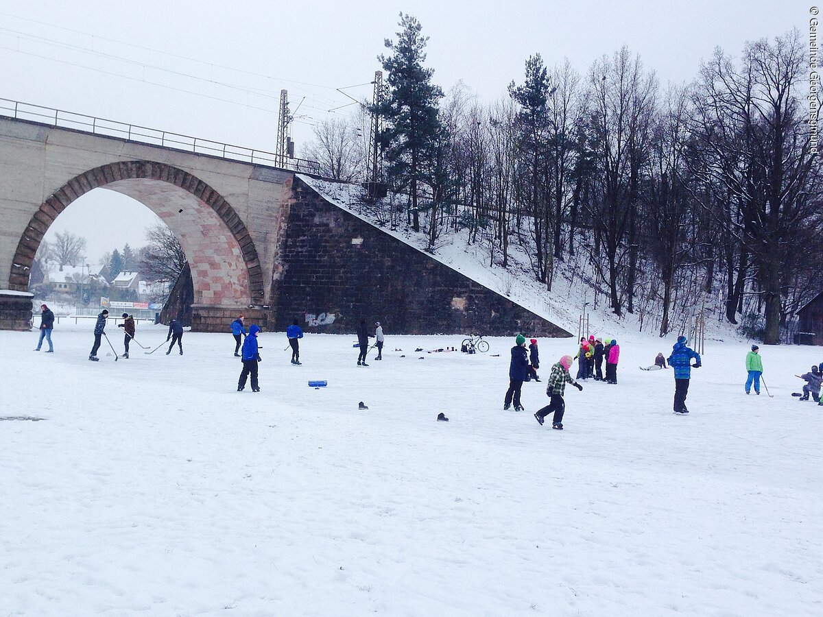 Ein Eislaufplatz mit vielen Eisläufern auf beschneiter Fläche. Im Hintergrund eine Bahnbrücke angrenzend ein verschneiter Hang mit kahlen Bäumen.