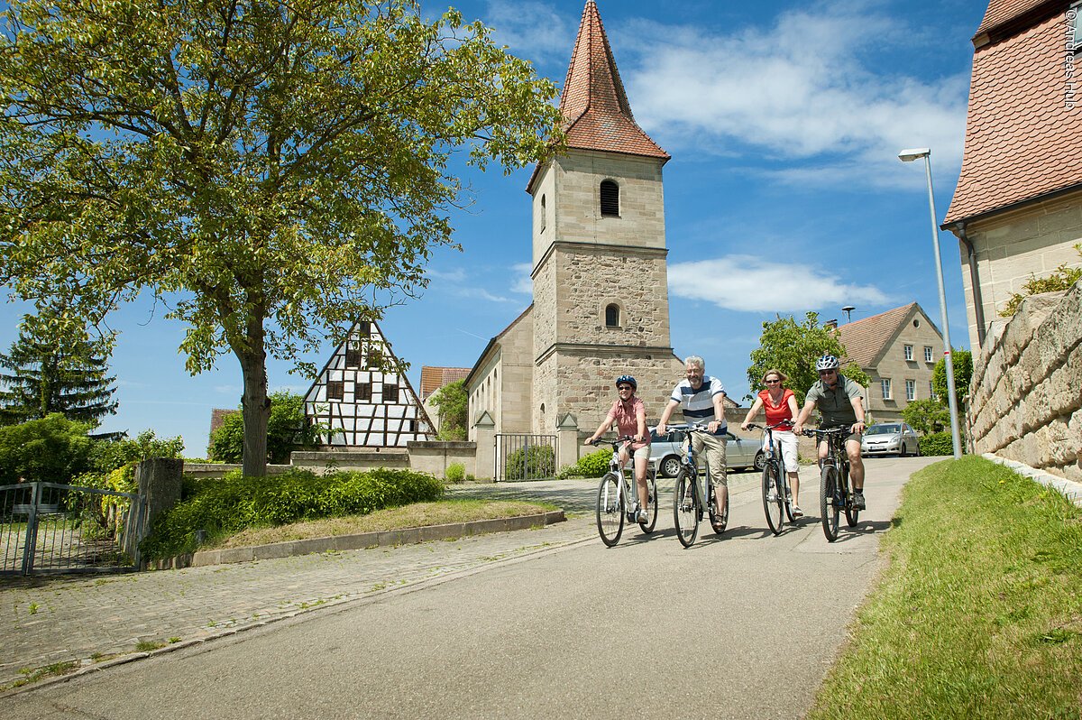 Zwei Männer und zwei Frauen fahren auf ihren Fahrrädern auf einer geteerten Straße durch ein Dorf. Im Hintergund stehen einzelne Häuser und ein großer Kirchturm.