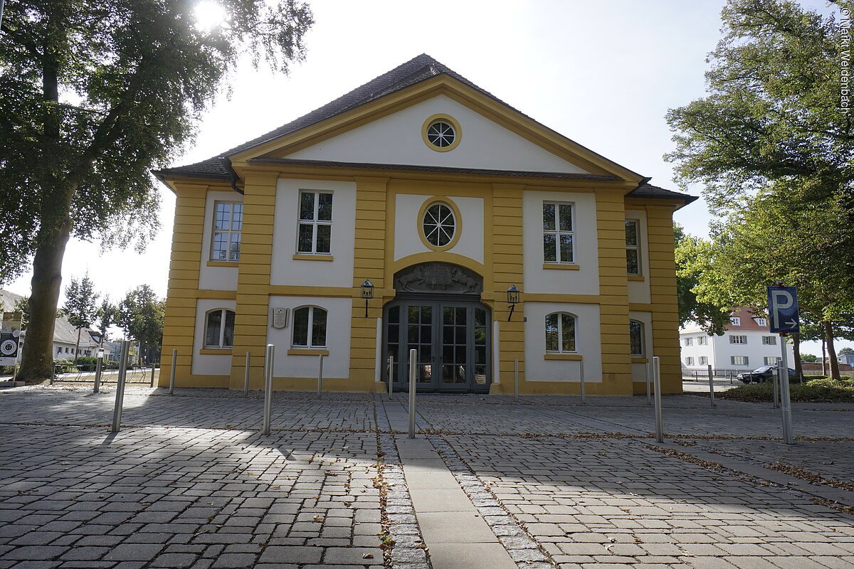 Ein barockes Haus mit zwei Ebenen. Die Fassade ist weiß mit gelben Elementen. Die Eigangstür ist doppeltürig mit kleinen Fenstern. Davor eine breite gepflasterte Straße.