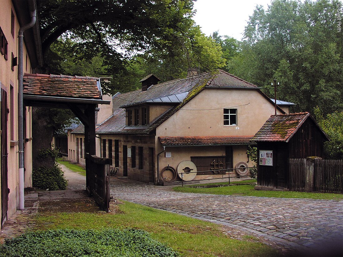 Eines der drei Museen der Stadt Roth, die der Museumsweg verbindet: Das Museum Historischer Eisenhammer Eckersmühlen. Ein altes Wohnhaus an welchem Mühlsteine lehnen. Daneben der Eingang eines weiteren alten Wohnhauses.