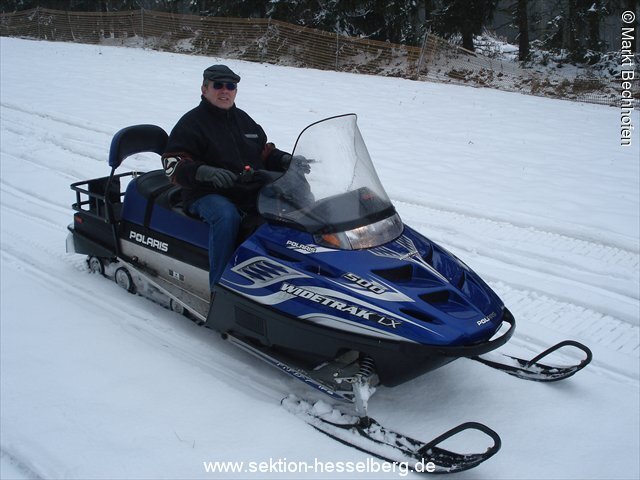 Ein Mann auf einem blauen Schneemobil lächelt in die Kamera. Er zieht eine Langlaufloipe durch den Schnee.