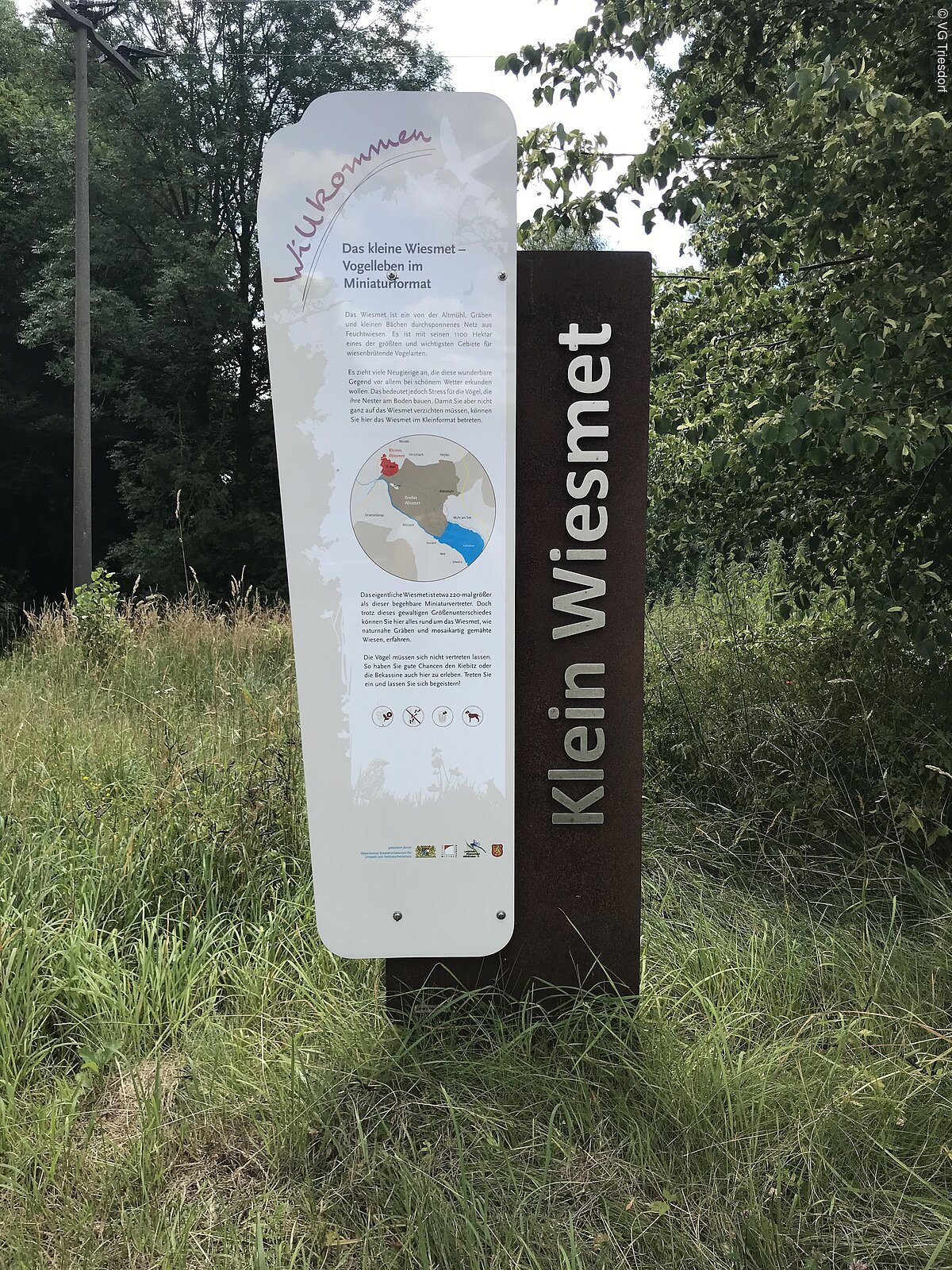 Eine Infotafel zu Klein Wiesmet steht in einer grünen Wiese, dahinter Bäume und Sträucher.