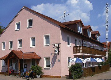 Ein dreistöckiger Gasthof mit roas farbender Fassade und rotem Ziegeldach. Entlang der östlichen Hauswand ein langer Balkon aus Holz. Darunter ein kleiner umzäunter Biergarten mit drei kleinen Sonnenschirmen.