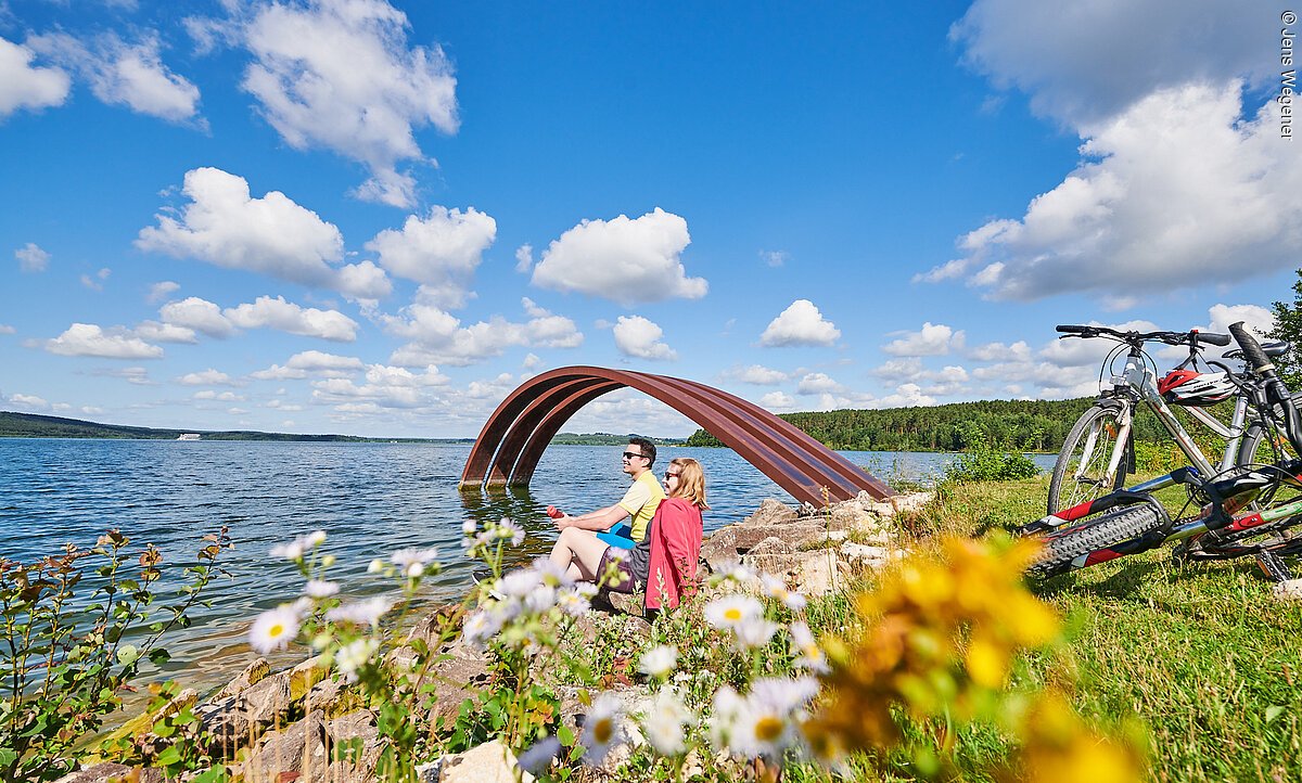 Ein junges Pärchen macht eine Pause vom Radeln am Ufer eines Sees. Sie sitzen direkt am Wasser auf einer grünen Wiese. Hinter ihnen stehen Fahrräder. Im Wasser steht ein roter Metallbogen.