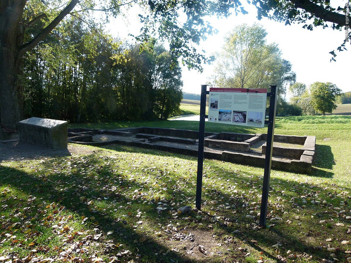 Eine römische Ruine auf einer grünen Wiese. Davor steht eine Infotafel.