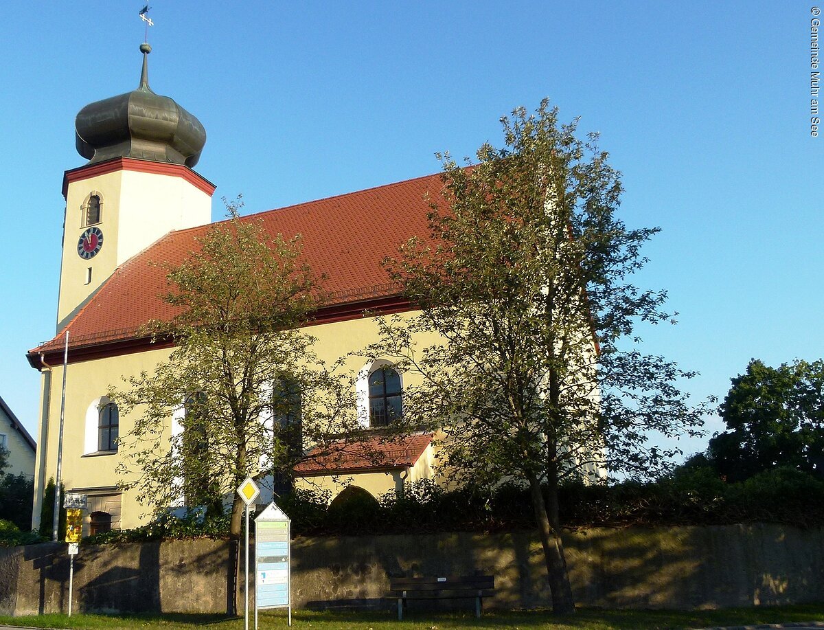 Ein kleine Kirche mit kurzem Kirchenschiff und kleinem Kirchturm mit Zwiebeldach. Die Kirche hat eine gelbe Fassade und ein rotes Ziegeldach. Vor der Kirche stehen zwei große Bäume und eine Steinmauer.