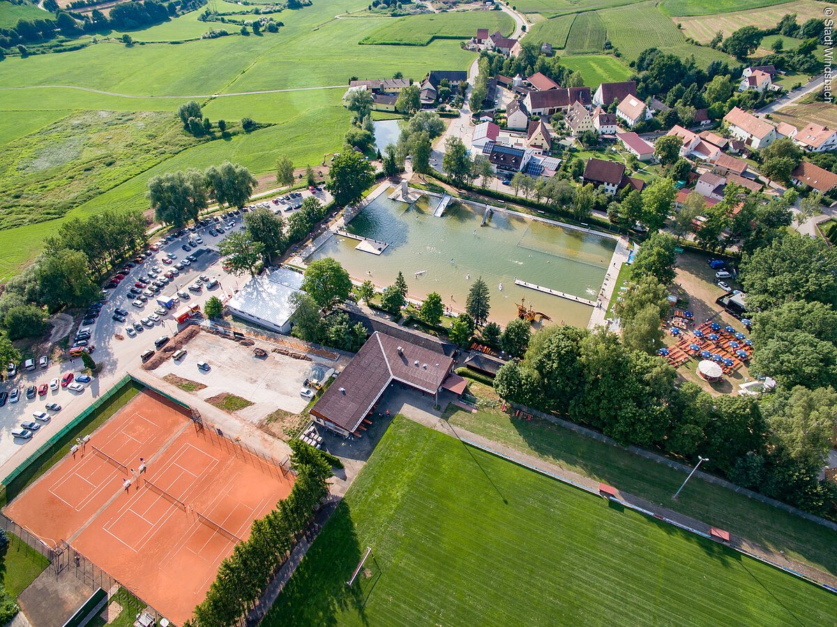 Luftbild eines Naturfreibads, links daneben ein roter Tennisplatz. Rechts neben dem Freibad eine Wohnsiedlung. Im Hintergrund erstrecken sich Felder.