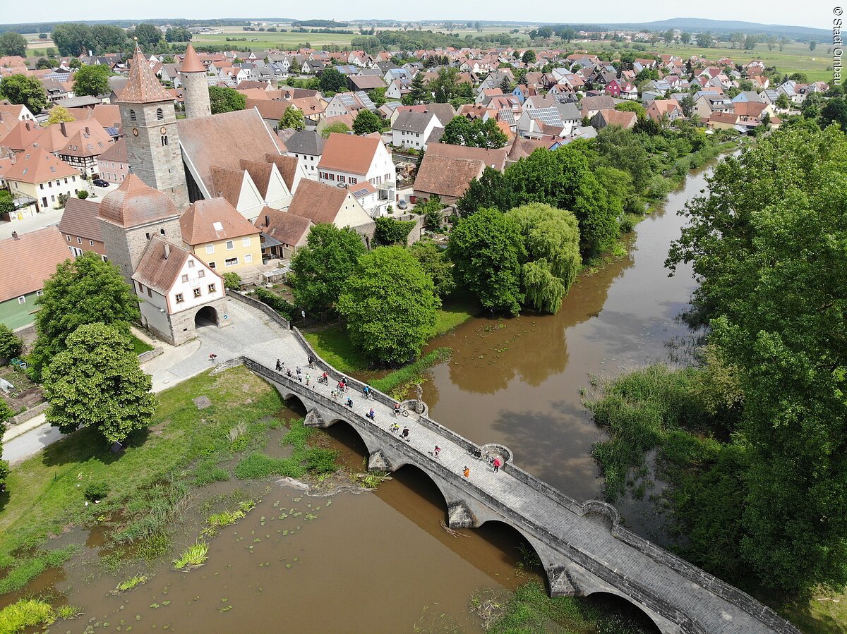 Luftbild der Stadt Ornbau. Eine lange Steinbrücke führt über einen überschwemmten Fluss zu einem Stadttor. Dahinter weitere Häuser einer Altstadt. Kleiner im Hintergund weitere Wohngebiete mit Häusern.